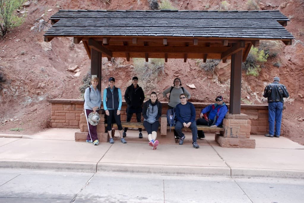 A group of seven people sitting under a wooden shelter on a roadside, with a rocky hillside in the background.