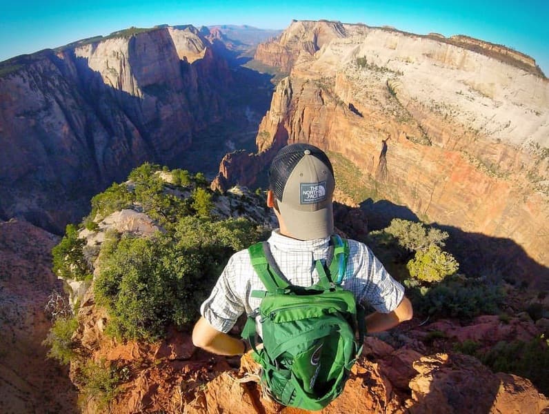 A person with a green backpack stands on a cliff overlooking a vast canyon landscape.