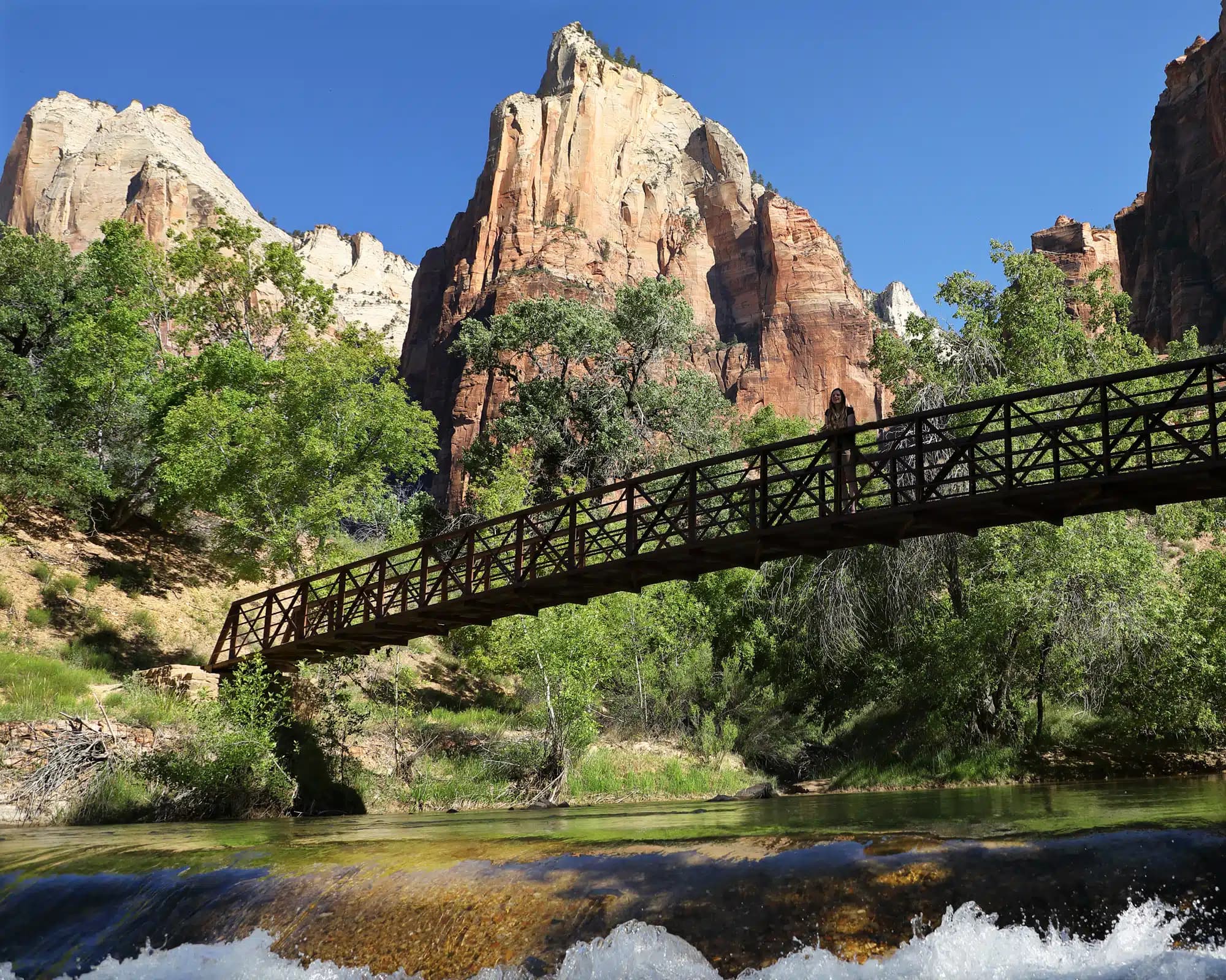 A wooden bridge spans over a stream surrounded by towering red cliffs and lush greenery.