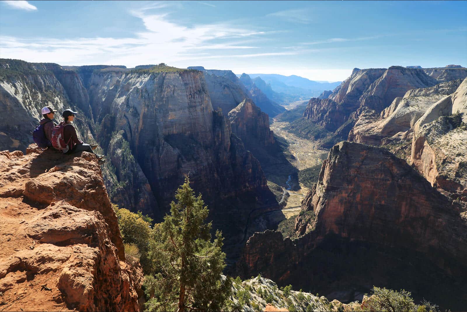 Two hikers sit on a cliff overlooking a vast canyon landscape.