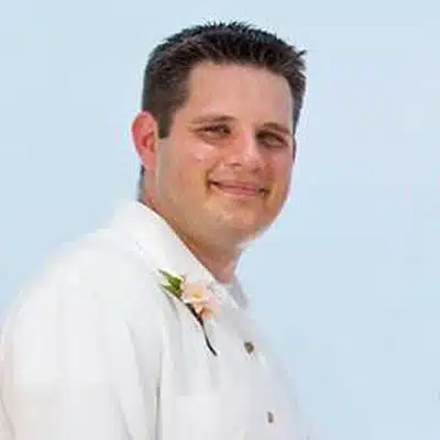 A man smiles while wearing a white shirt with a flower boutonnière.