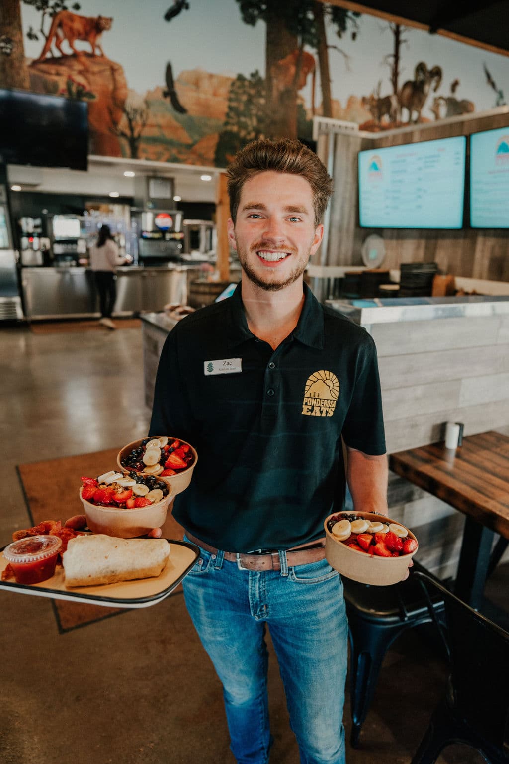 A smiling server holds a tray of colorful food and two bowls in a casual dining setting.
