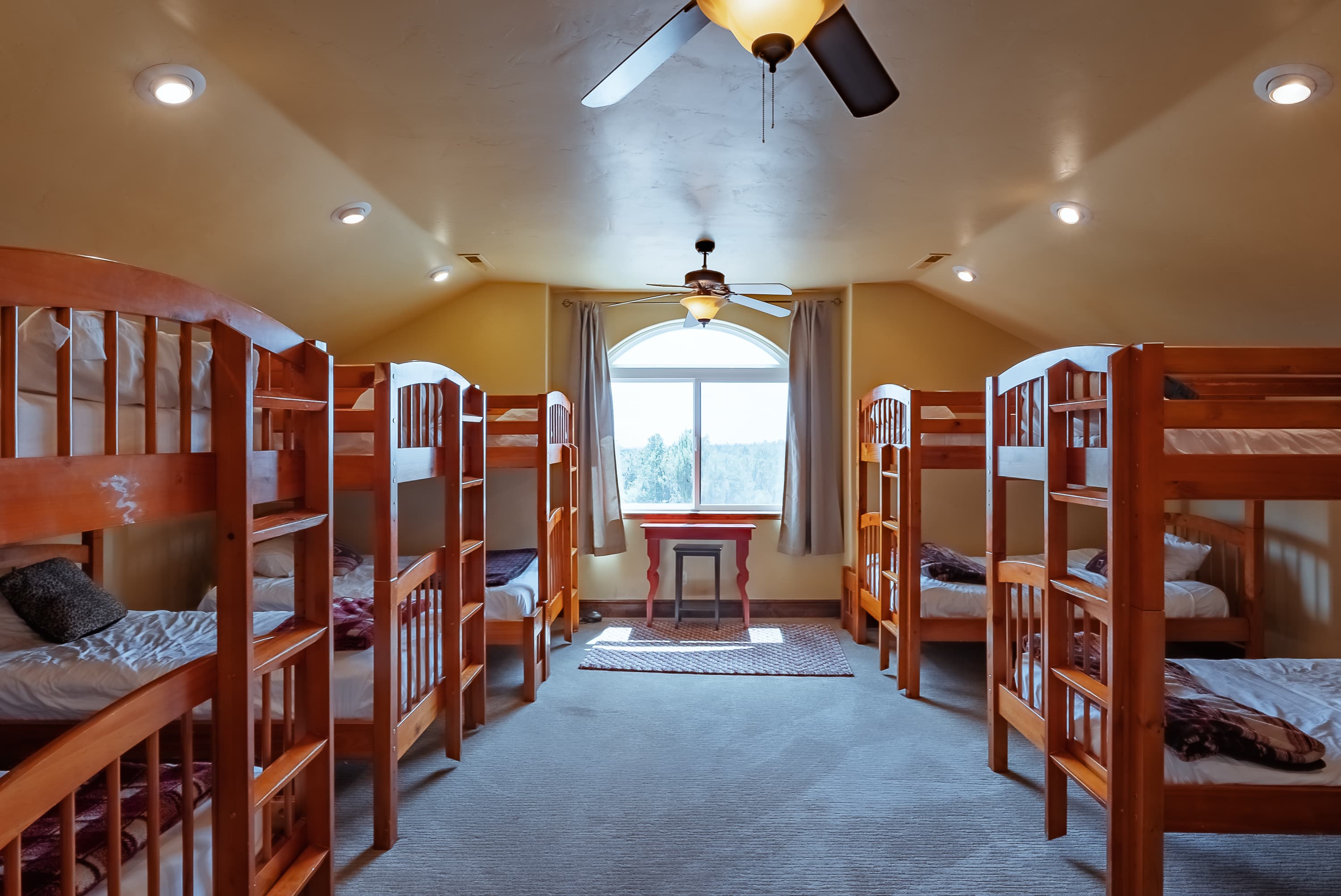 A spacious attic room with two sets of wooden bunk beds, a small table, and a large window letting in natural light.