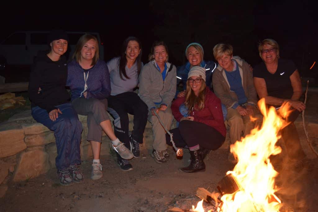 A group of eight women sits around a campfire at night, smiling and enjoying each other's company.