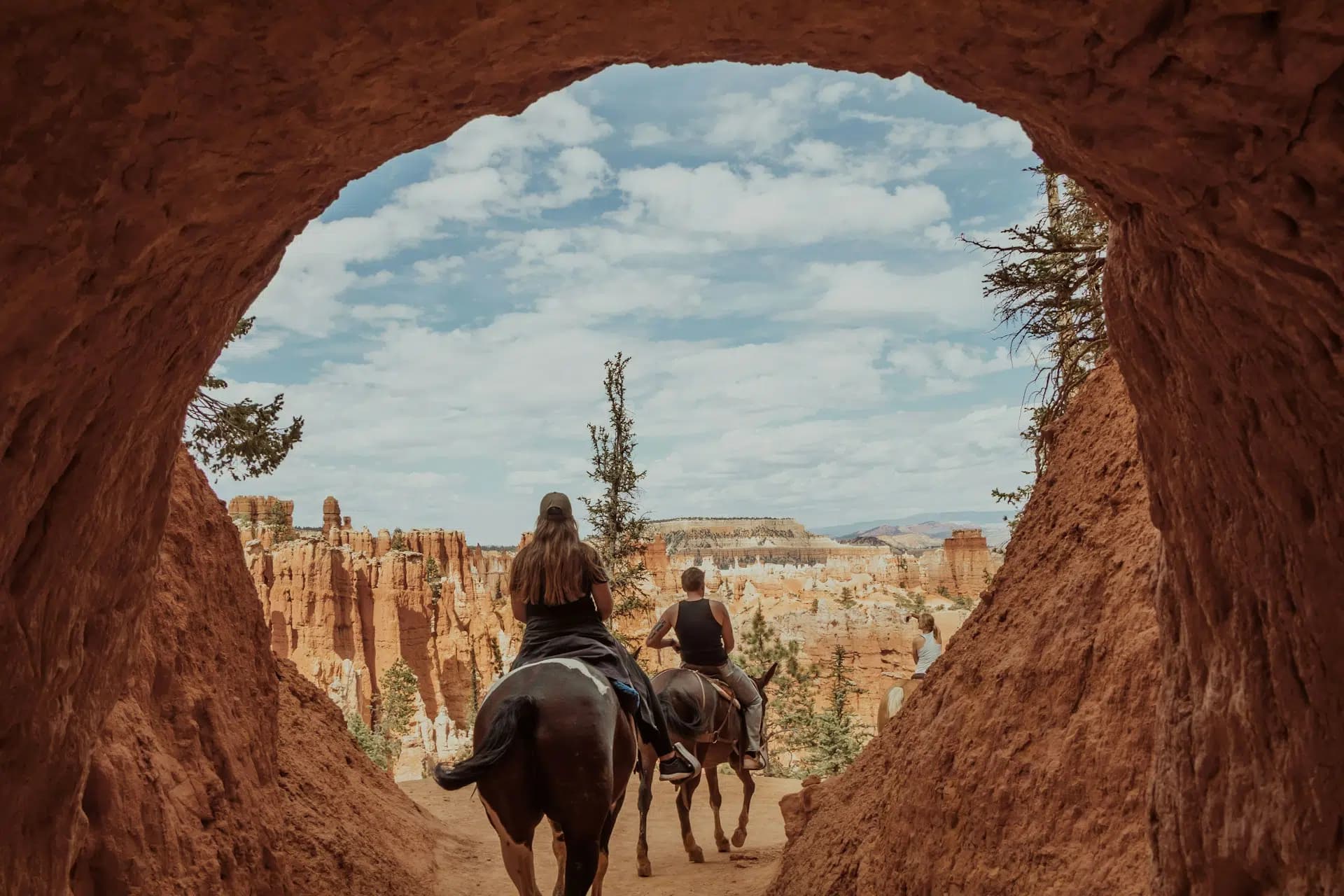 Two people on horseback ride through a rock archway with a scenic landscape behind them.