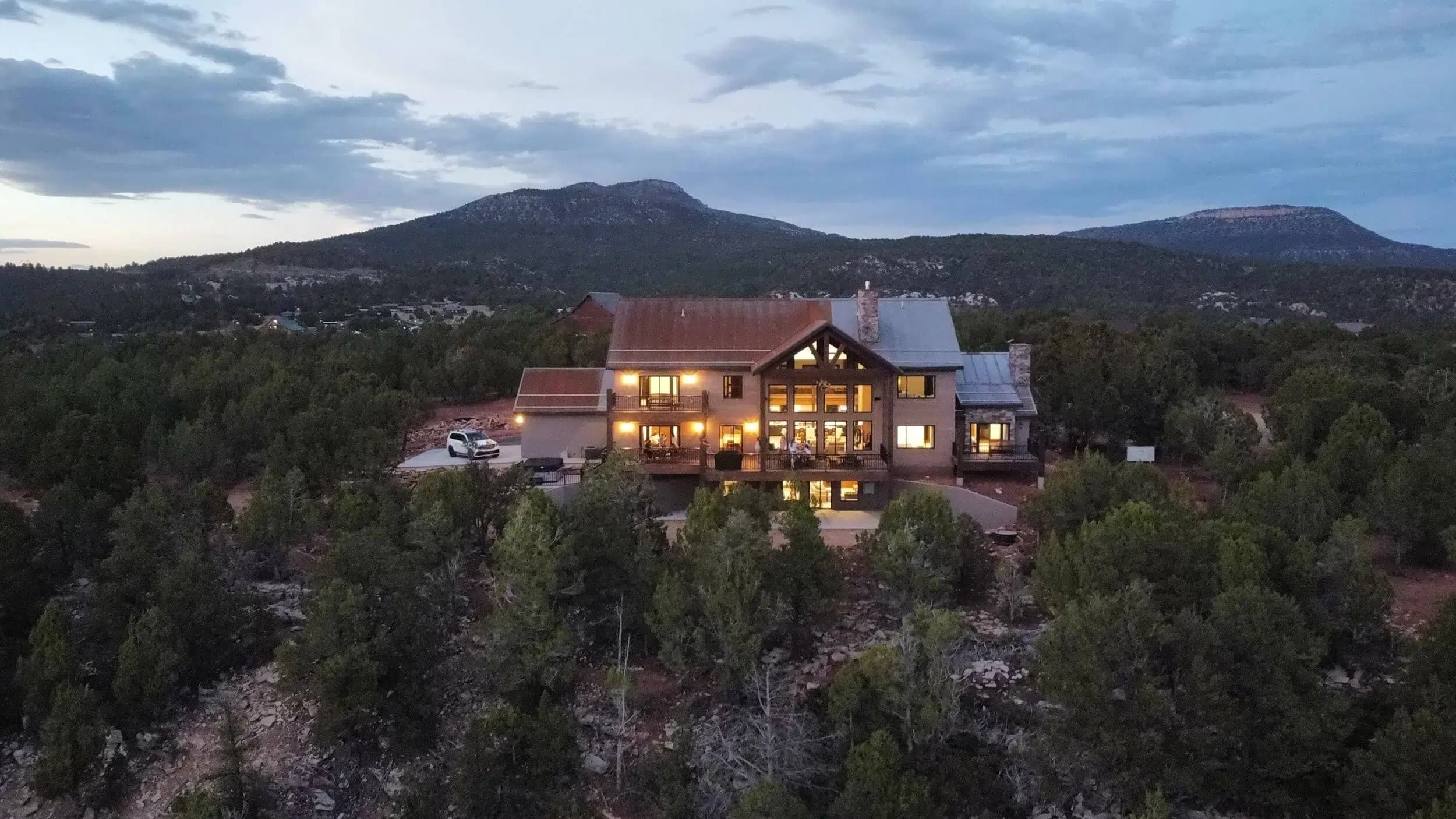 A large house with warm lights is nestled among trees, set against a backdrop of mountains under a cloudy sky.