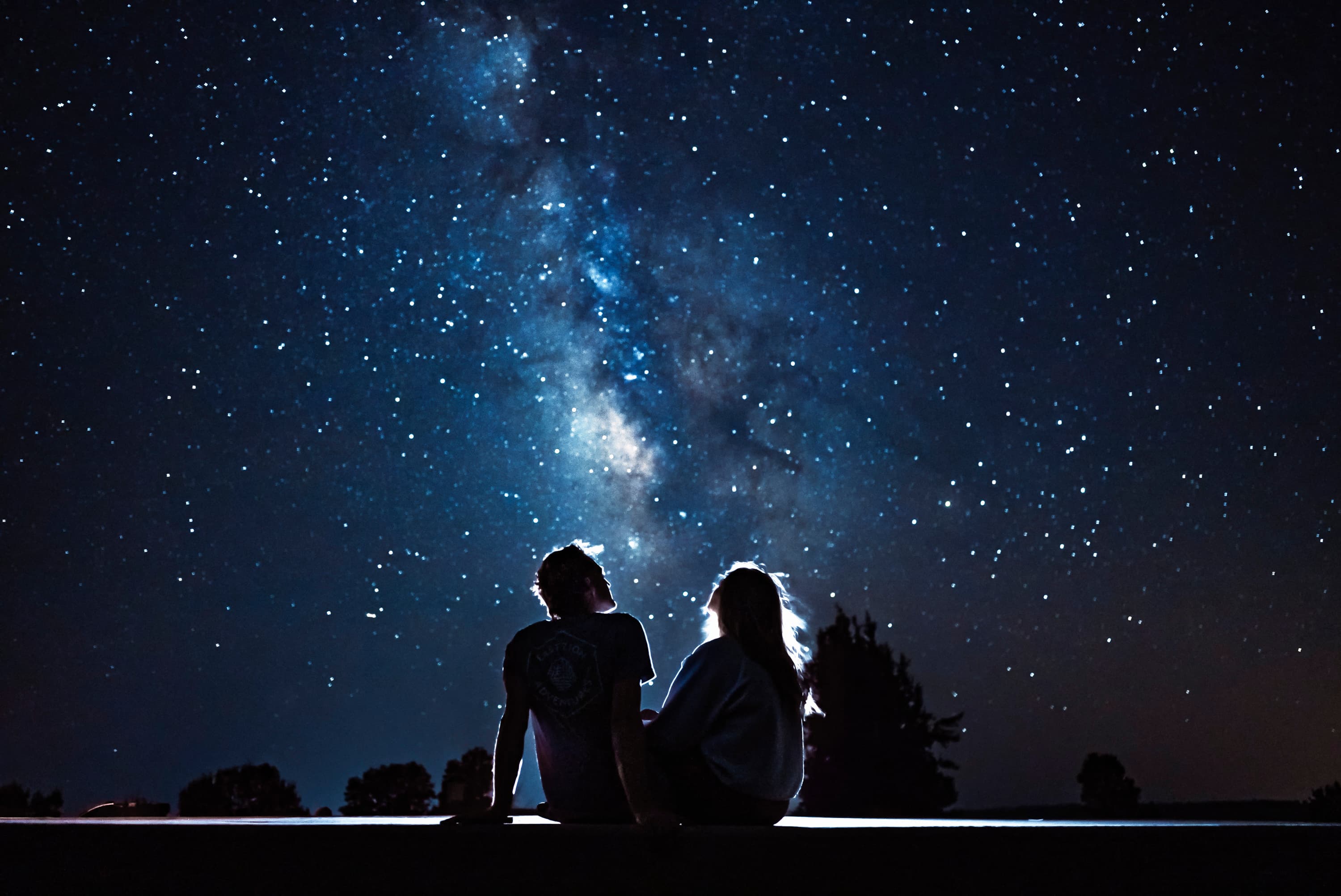 A couple sits together looking up at a star-filled night sky.