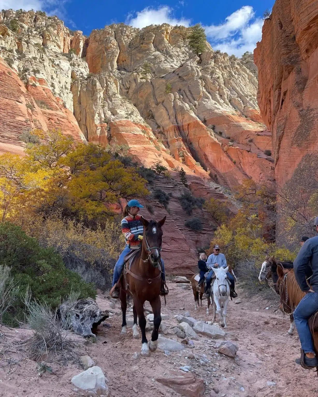 Four riders on horseback navigate a rocky trail surrounded by vibrant red cliffs and autumn foliage.