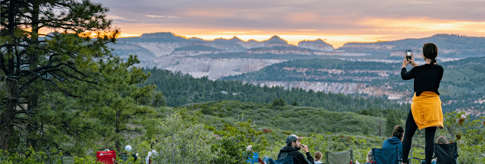A person takes a photo of a colorful sunset over a mountainous landscape while seated campers enjoy the view.