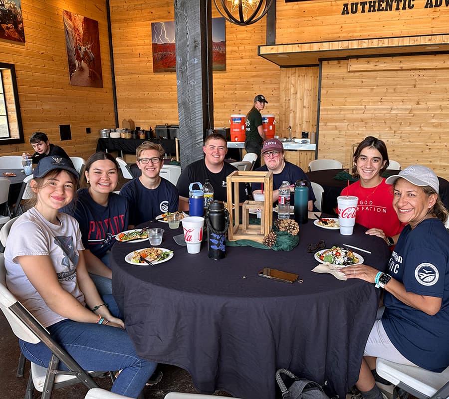 A group of six people enjoy a meal together at a round table in a rustic dining space.