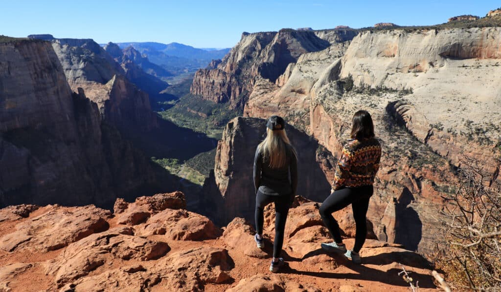 Two women stand on a rocky ledge overlooking a vast canyon landscape.