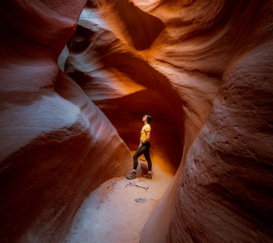 A person stands in a narrow, brightly lit canyon with smooth, reddish rock formations.