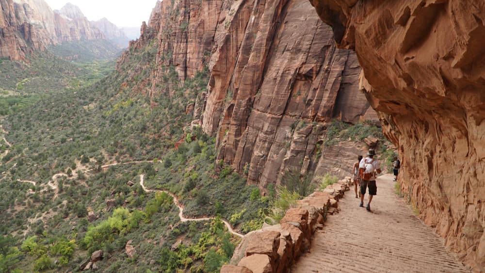 Hikers walking along a narrow path surrounded by steep rock formations and lush greenery in a canyon.