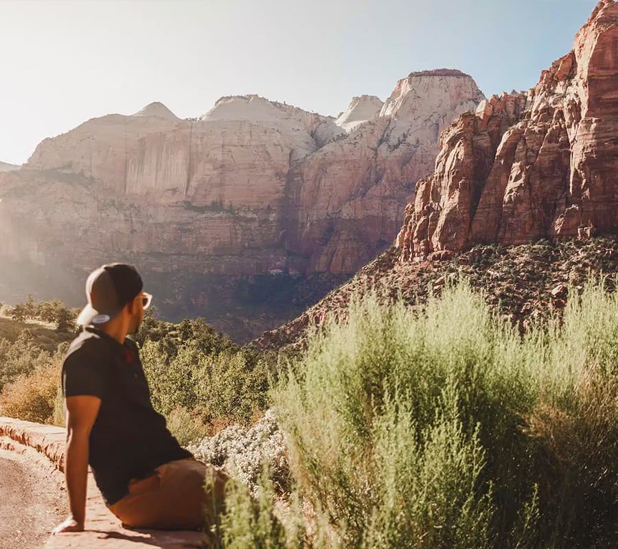 A person sits on a rocky outcrop, gazing at towering red cliffs in a sunny landscape.