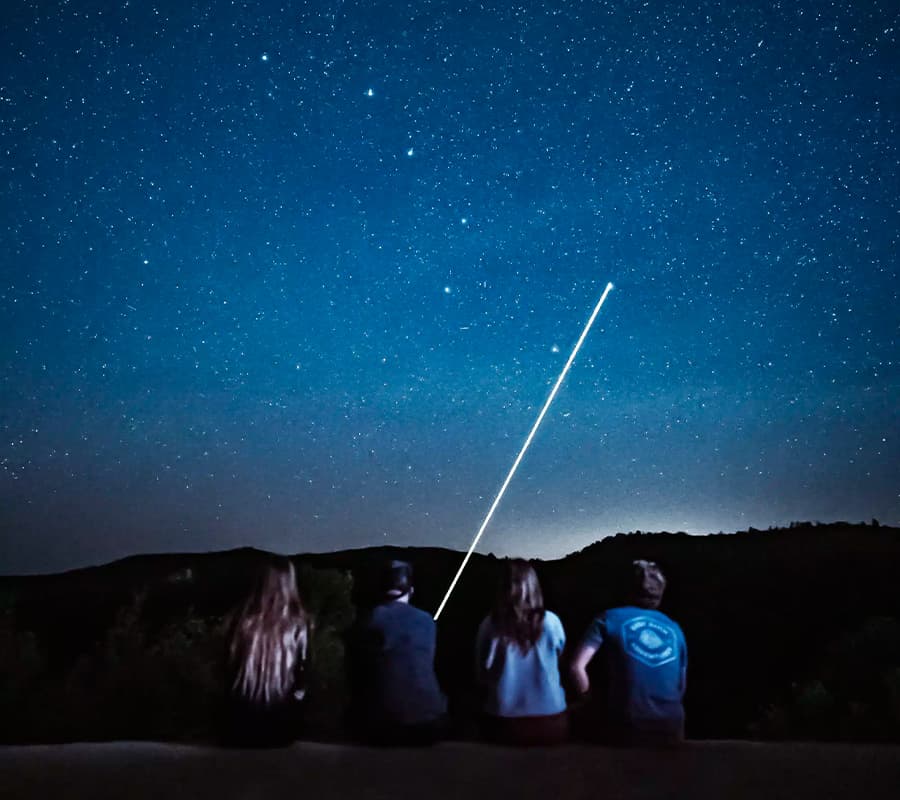 Four people sit on a ledge gazing up at a starry night sky with a bright streak of light.