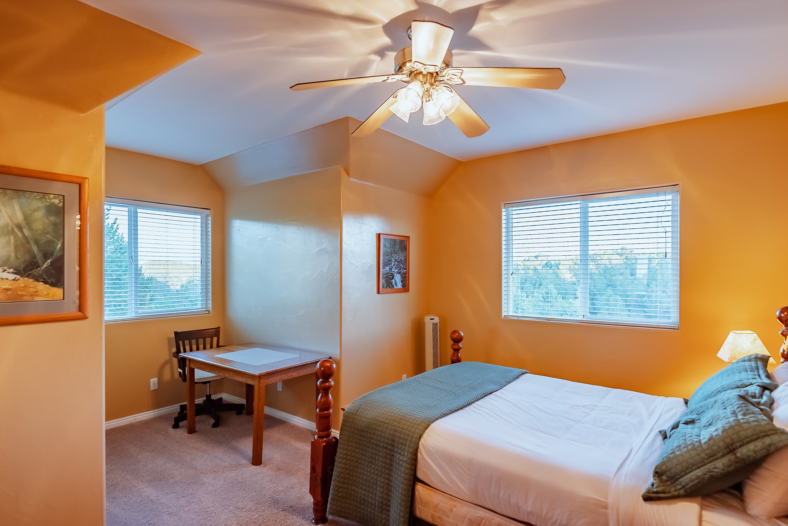 Cozy bedroom with a bed, desk, and large windows, featuring warm orange walls and a ceiling fan.