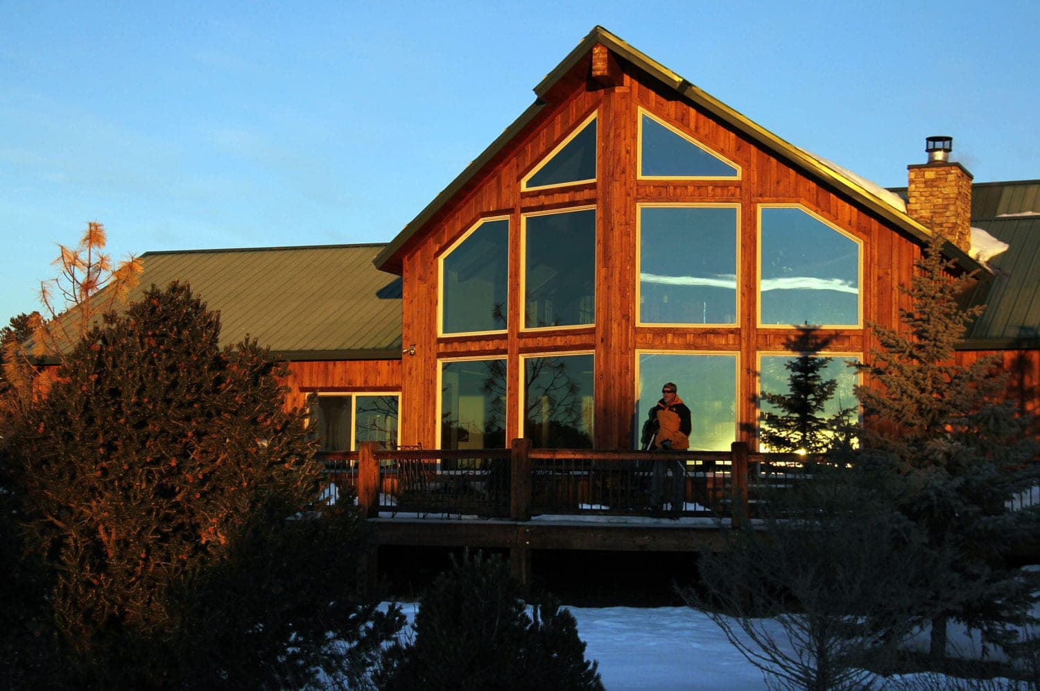 A wooden house with large windows and a person standing on the deck during sunset.
