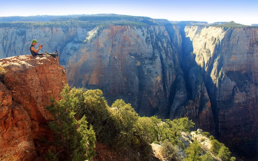 A person sits on a rock ledge overlooking a deep canyon surrounded by rugged cliffs and greenery.