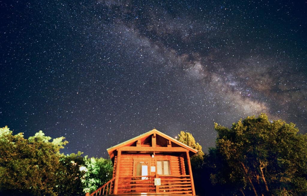 A wooden cabin sits beneath a star-filled sky with the Milky Way visible above.