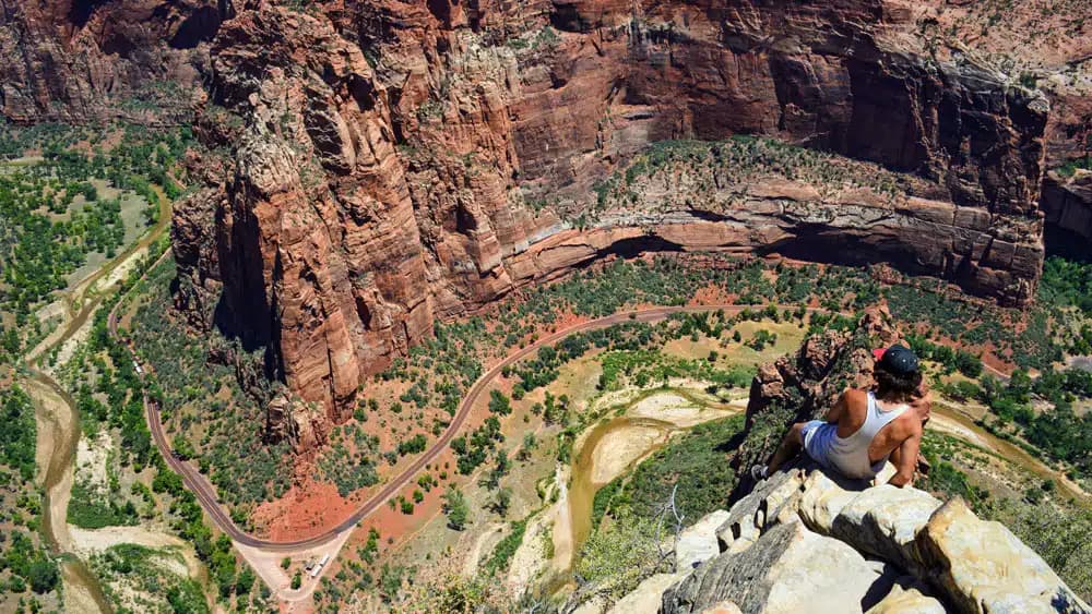 A person sits on a rocky ledge overlooking a winding river through a vast canyon.