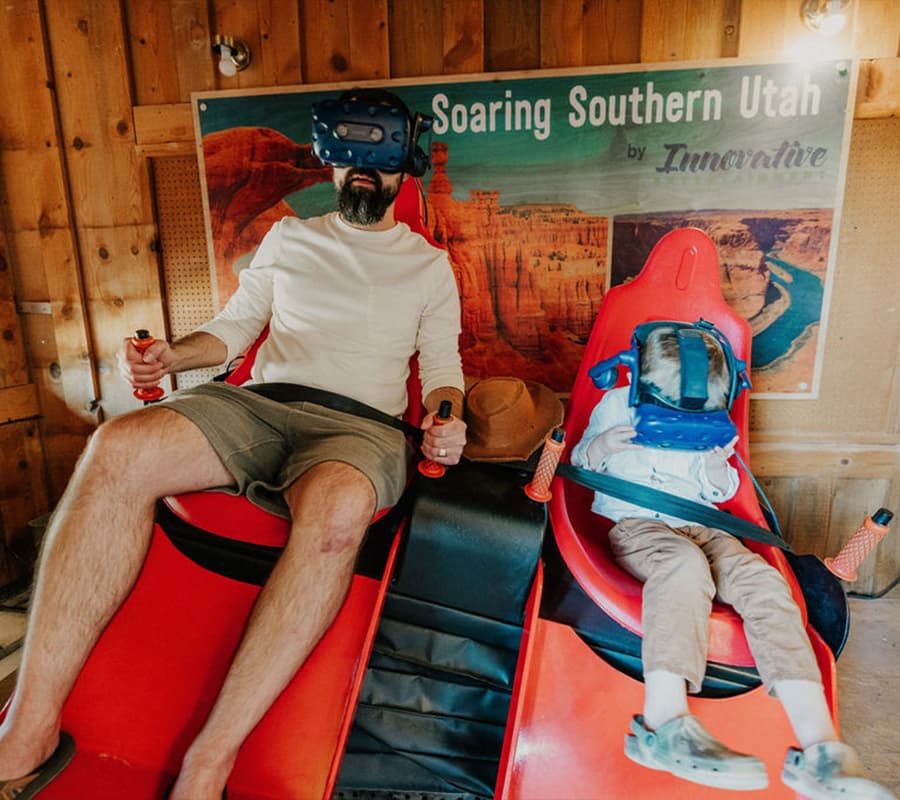 A man and a child both wearing VR headsets sit on red seats with a scenic poster of Southern Utah in the background.