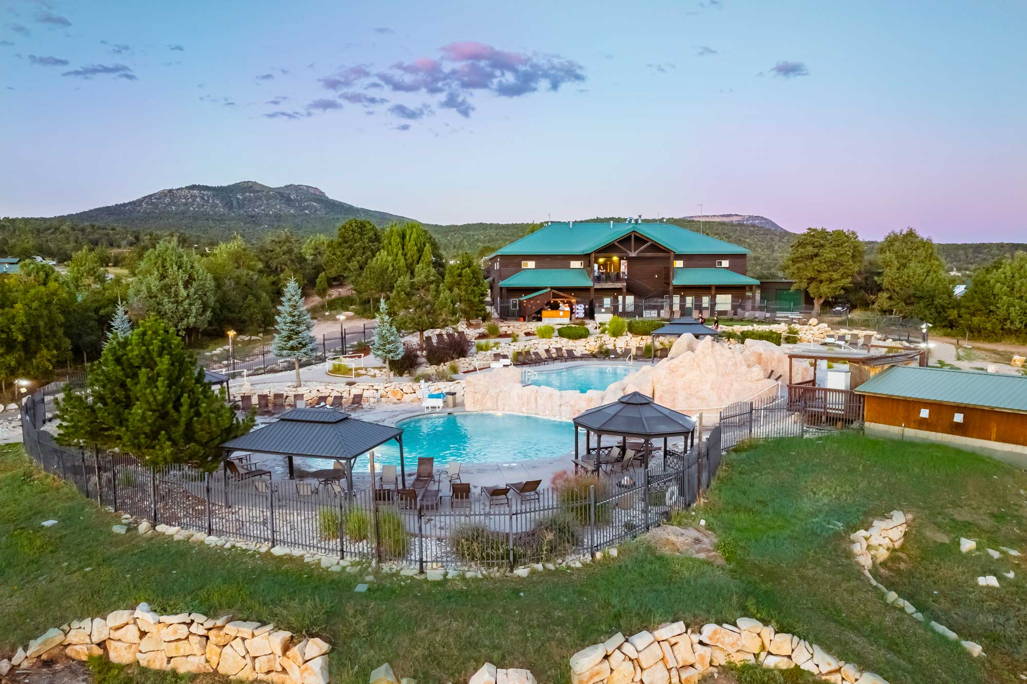 Aerial view of a resort with a pool, surrounded by greenery and mountains under a twilight sky.
