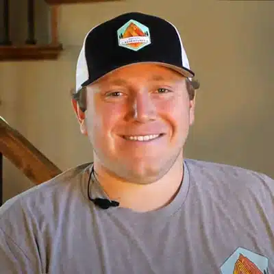 A smiling young man wearing a baseball cap and microphone sits in a cozy indoor setting.