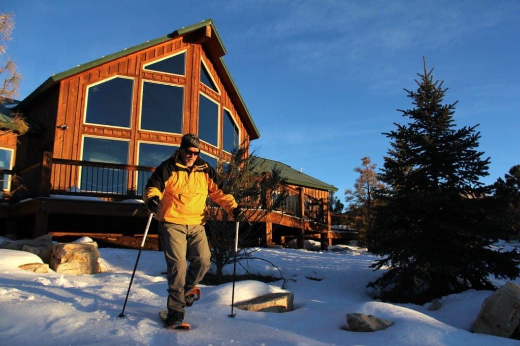 A person in a yellow jacket is snowshoeing in front of a wooden cabin surrounded by snow.