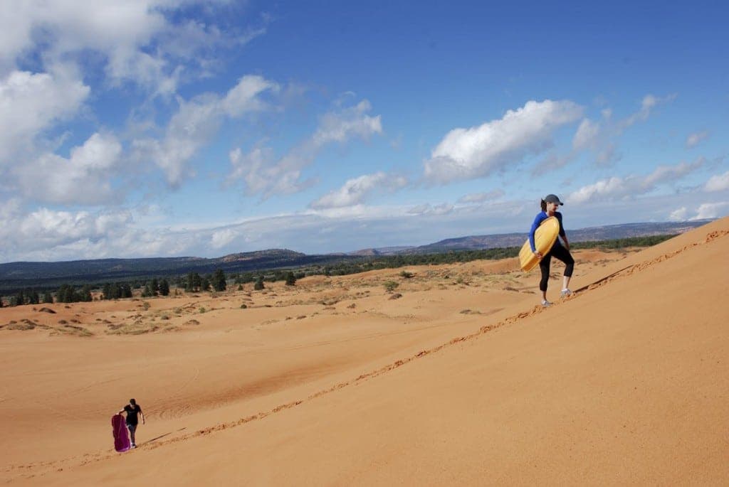 A person carries a surfboard while climbing a sandy dune, with another individual walking in the background.