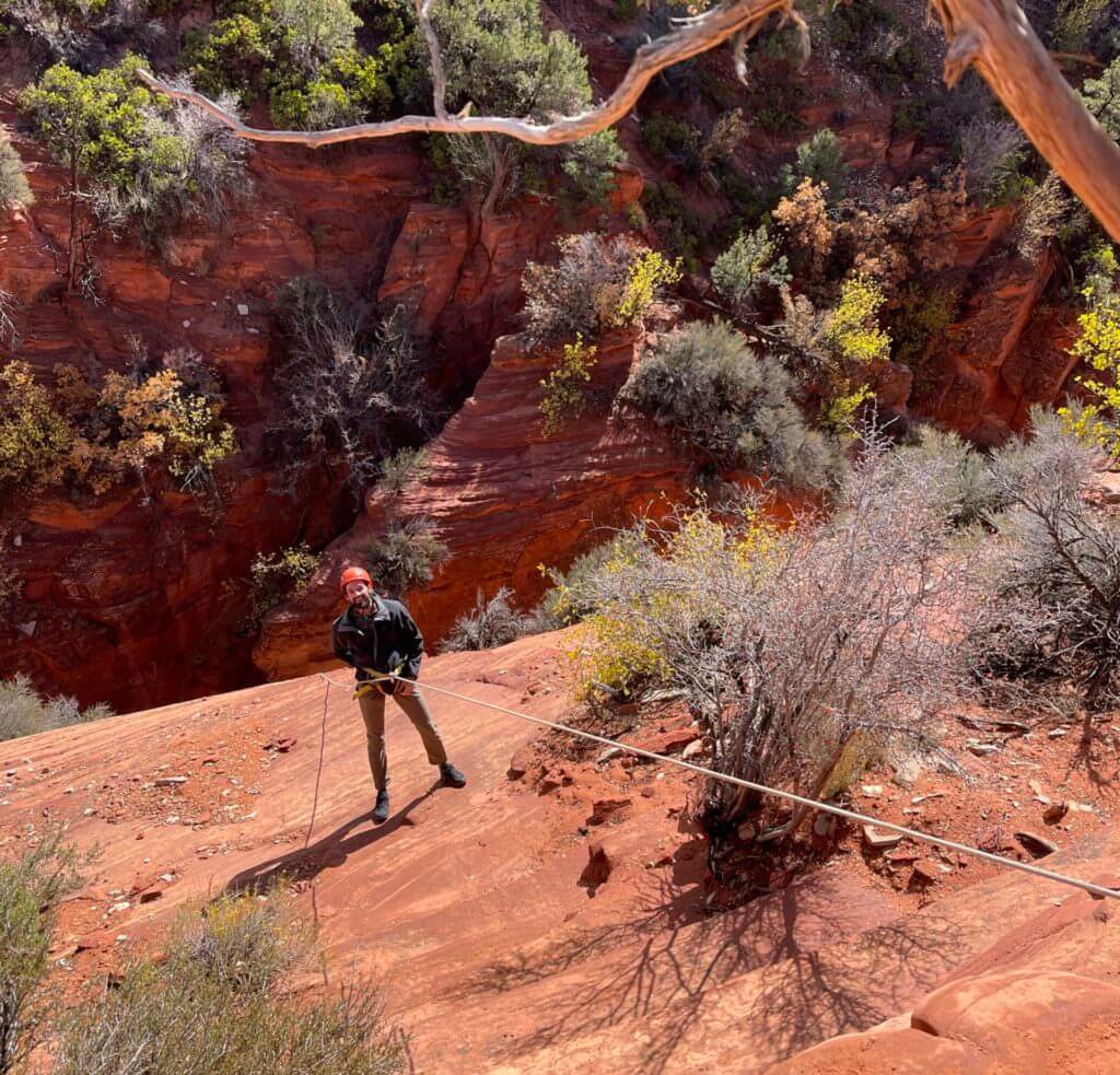A person rappels down a steep red rock slope with greenery in the background.