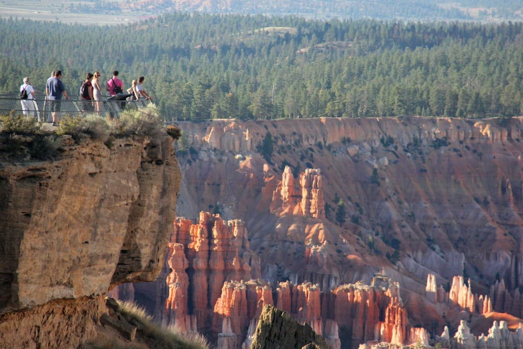 Visitors stand on a cliff overlooking colorful rock formations and a forested valley.