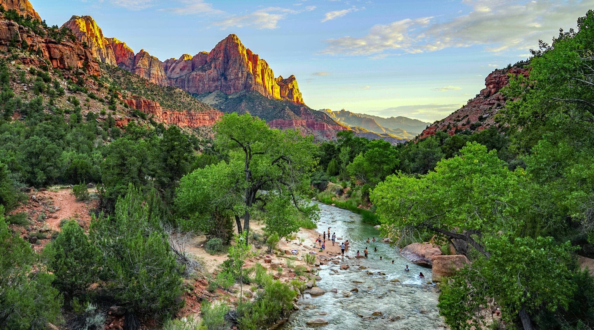 A serene river flows through a lush green valley surrounded by majestic mountains under a colorful sky.