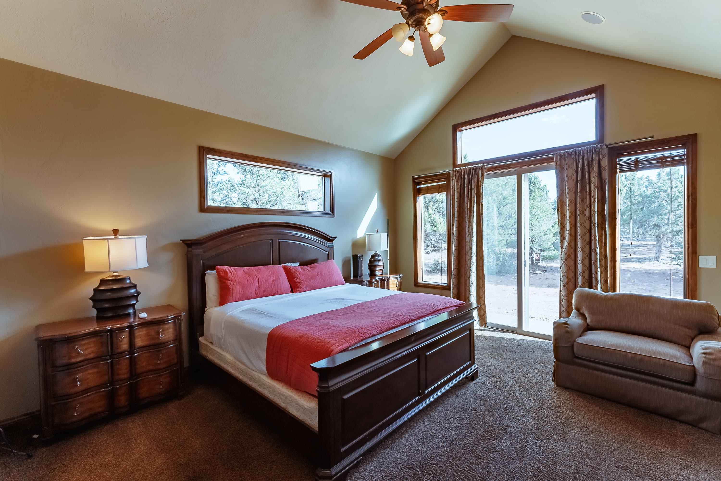 A cozy bedroom featuring a queen bed with red accents, a wooden dresser, and large windows overlooking a natural landscape.