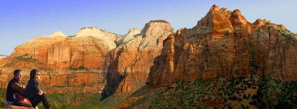 A couple sits on a ledge overlooking towering red rock formations at Zion National Park.