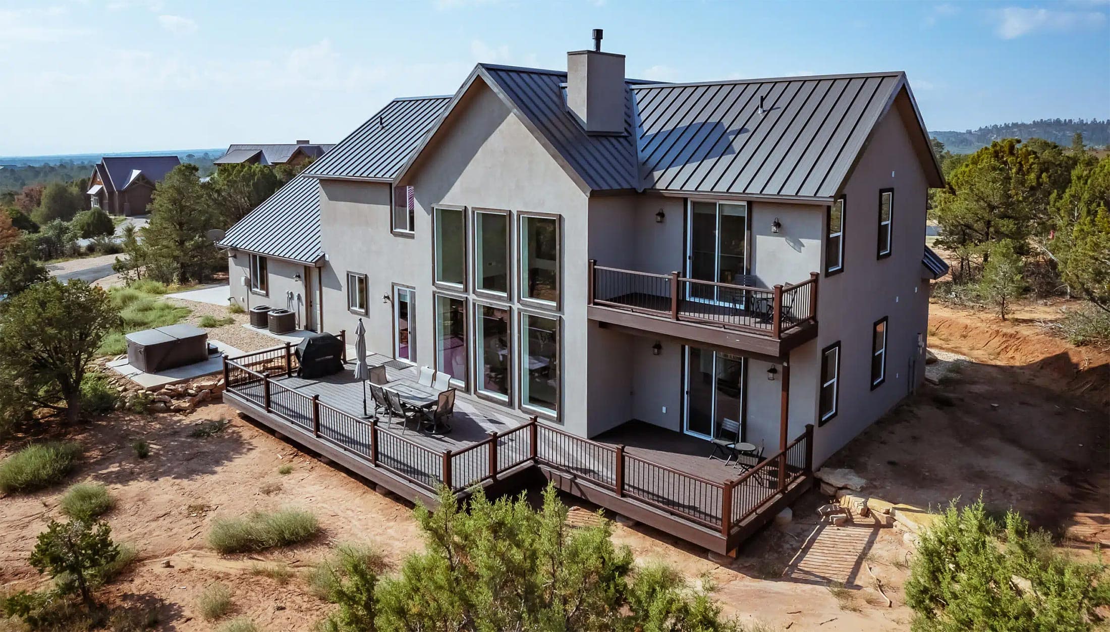 A modern two-story house with large windows and a wooden deck, surrounded by greenery and outdoor space.