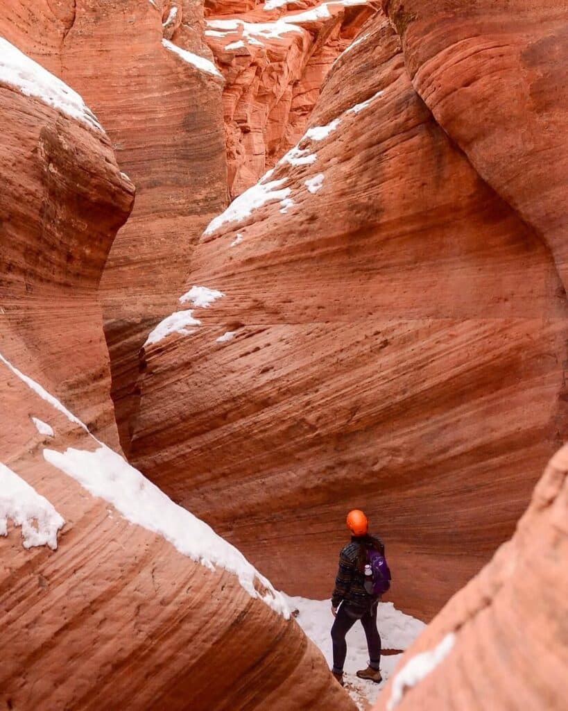 A person wearing an orange helmet stands in a narrow, rocky canyon with red and brown swirled formations.
