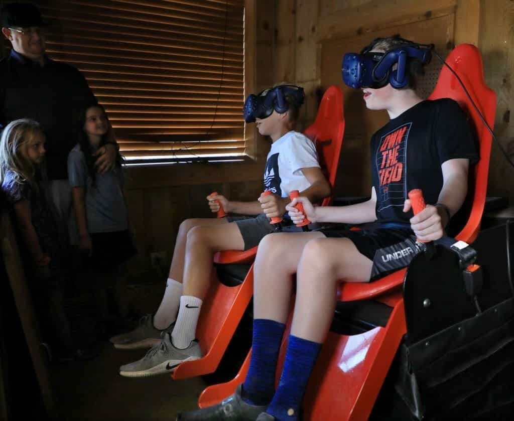 Two boys with virtual reality headsets sit in red chairs while two girls and an adult watch them.