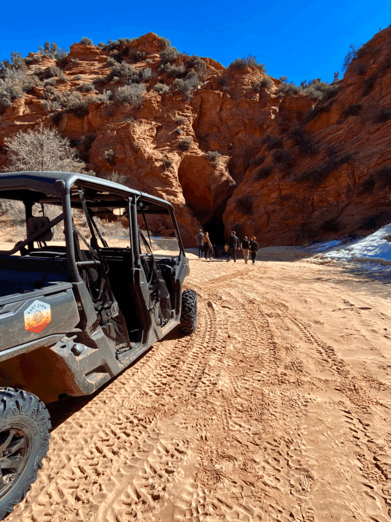 A rugged off-road vehicle parked on sandy terrain with a group of people heading towards a rocky cave entrance.