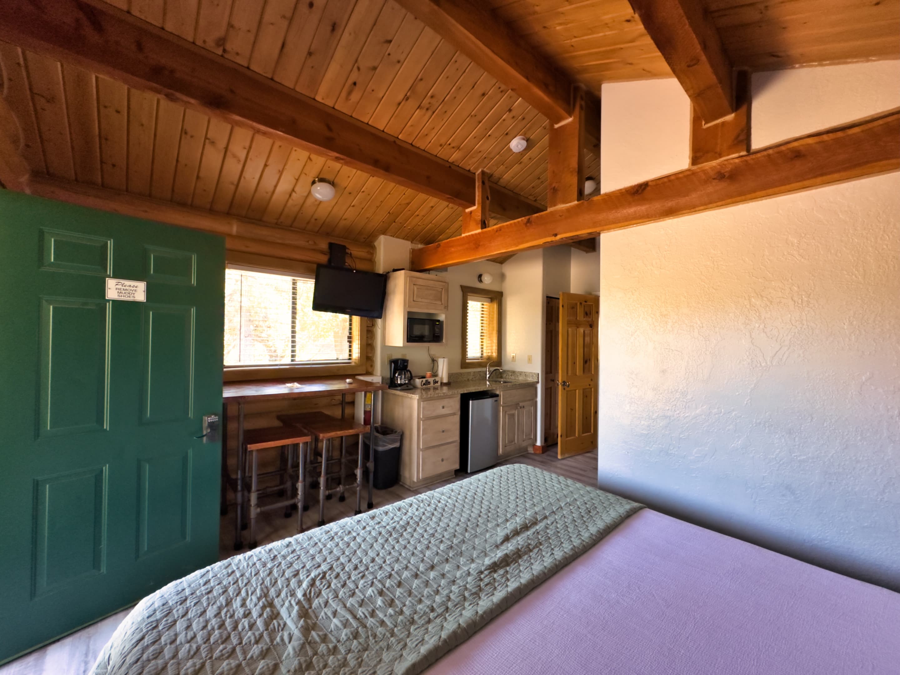 A cozy wooden cabin interior with a green door, a bed, kitchenette, and a small dining area.