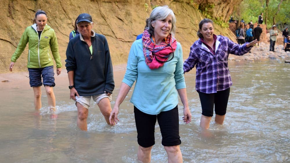 Four women walking through shallow water in a scenic outdoor setting.
