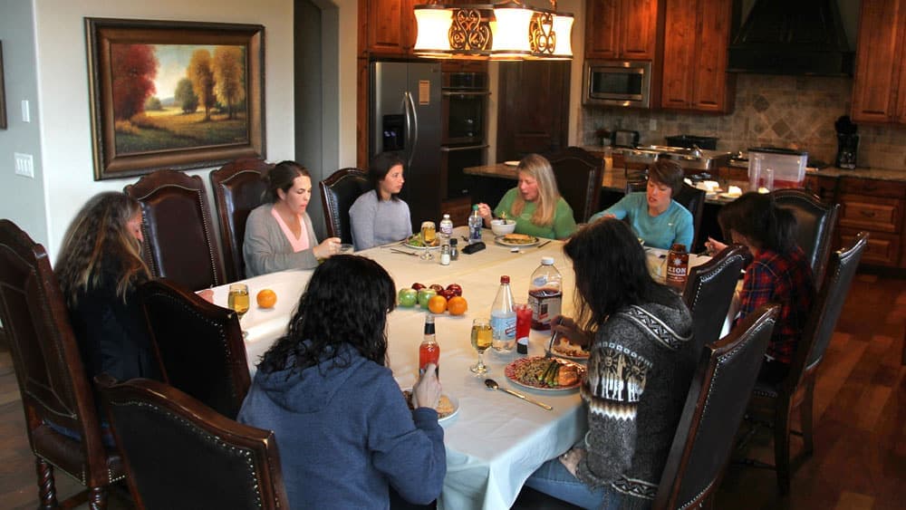 A group of eight women seated around a dining table, enjoying a meal in a warmly lit kitchen.