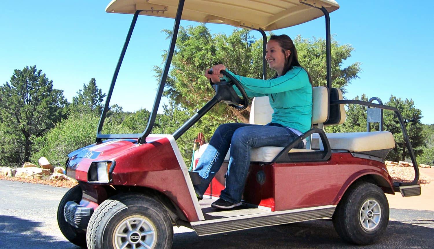 A woman smiles while driving a red golf cart on a sunny day surrounded by greenery.