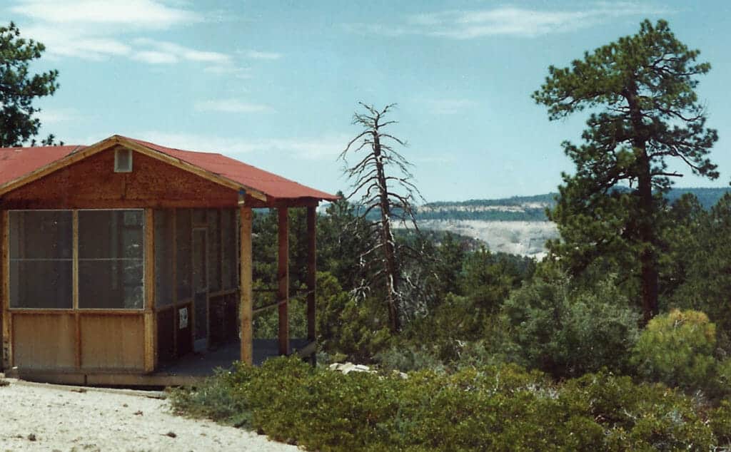 A wooden cabin surrounded by trees overlooks a distant landscape under a clear blue sky.