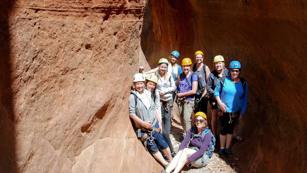 A group of eight climbers poses together in a narrow canyon, wearing helmets and harnesses.