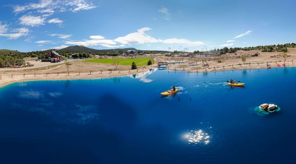 Two kayakers paddle on a bright blue lake surrounded by open land and a clear sky.