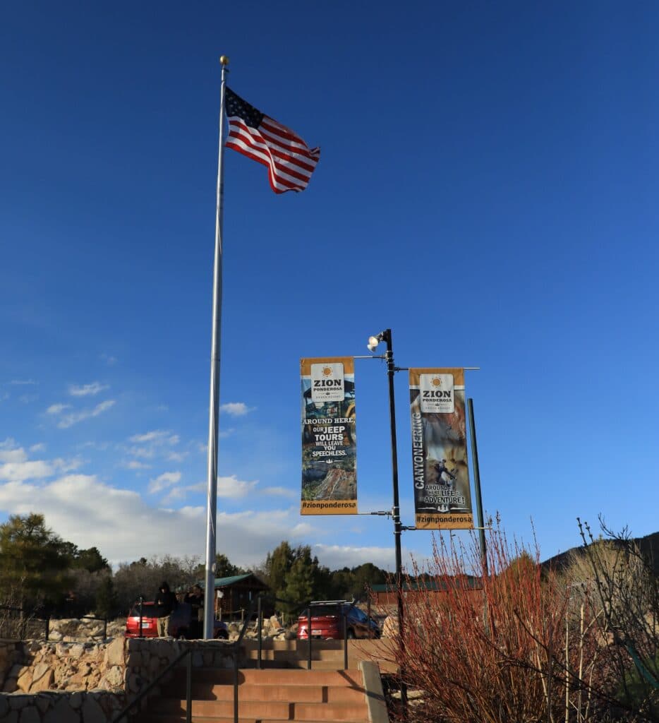 A U.S. flag flies beside informational banners at the entrance of Zion National Park against a blue sky.