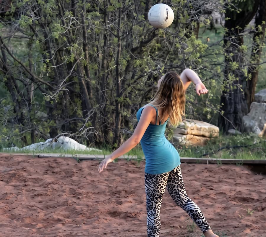 A woman wearing a blue tank top and leopard-print leggings is about to hit a volleyball in a sandy area surrounded by greenery.