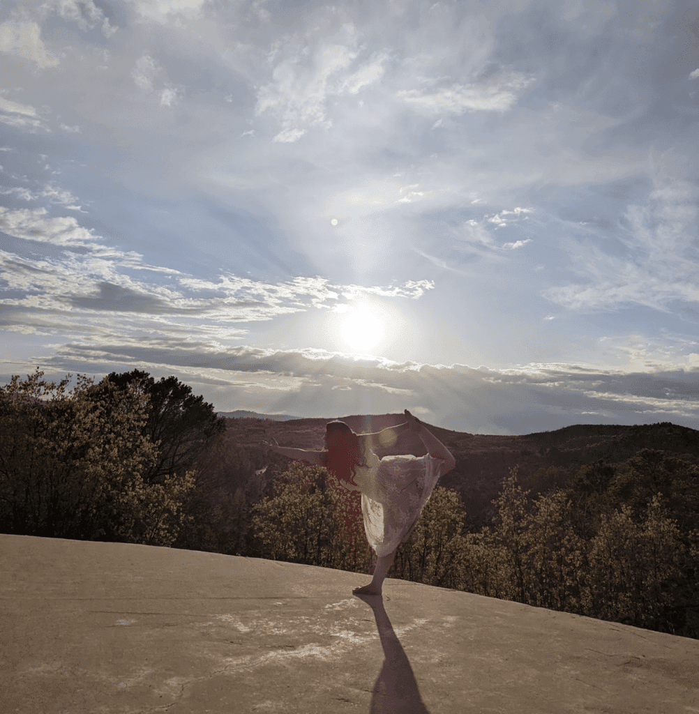 A person poses in a yoga stance on a ledge against a backdrop of a sunset and cloudy sky.