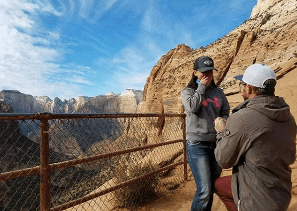 A man kneels to propose to a woman on a scenic overlook with mountains in the background.