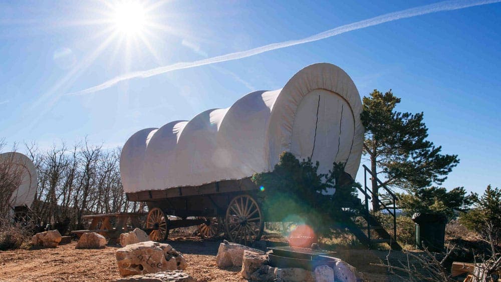A vintage covered wagon sits under a bright sun, surrounded by trees and rocks.